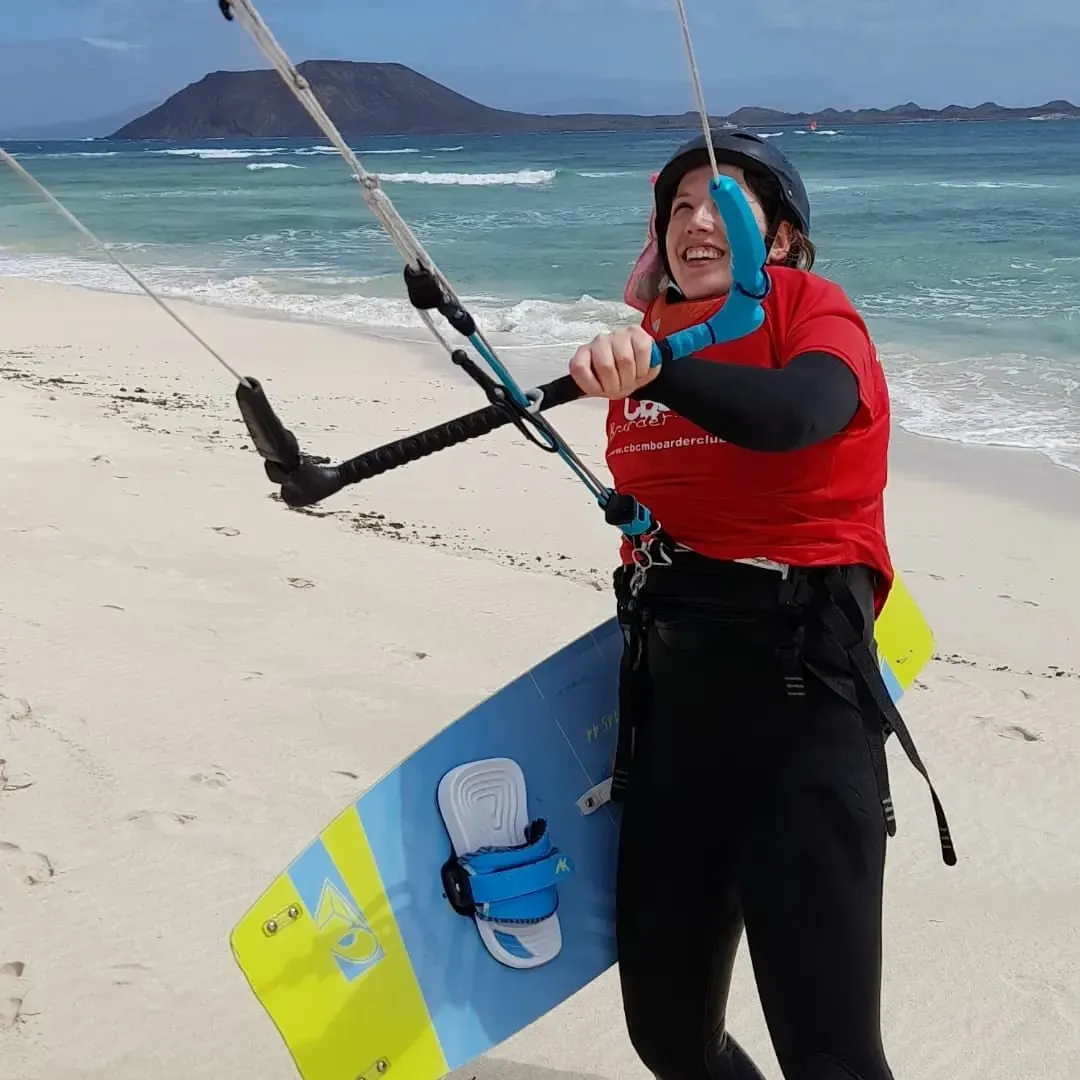 Alumna de CBCM sonriendo en la playa, con el arnés puesto, la barra de la cometa y la tabla