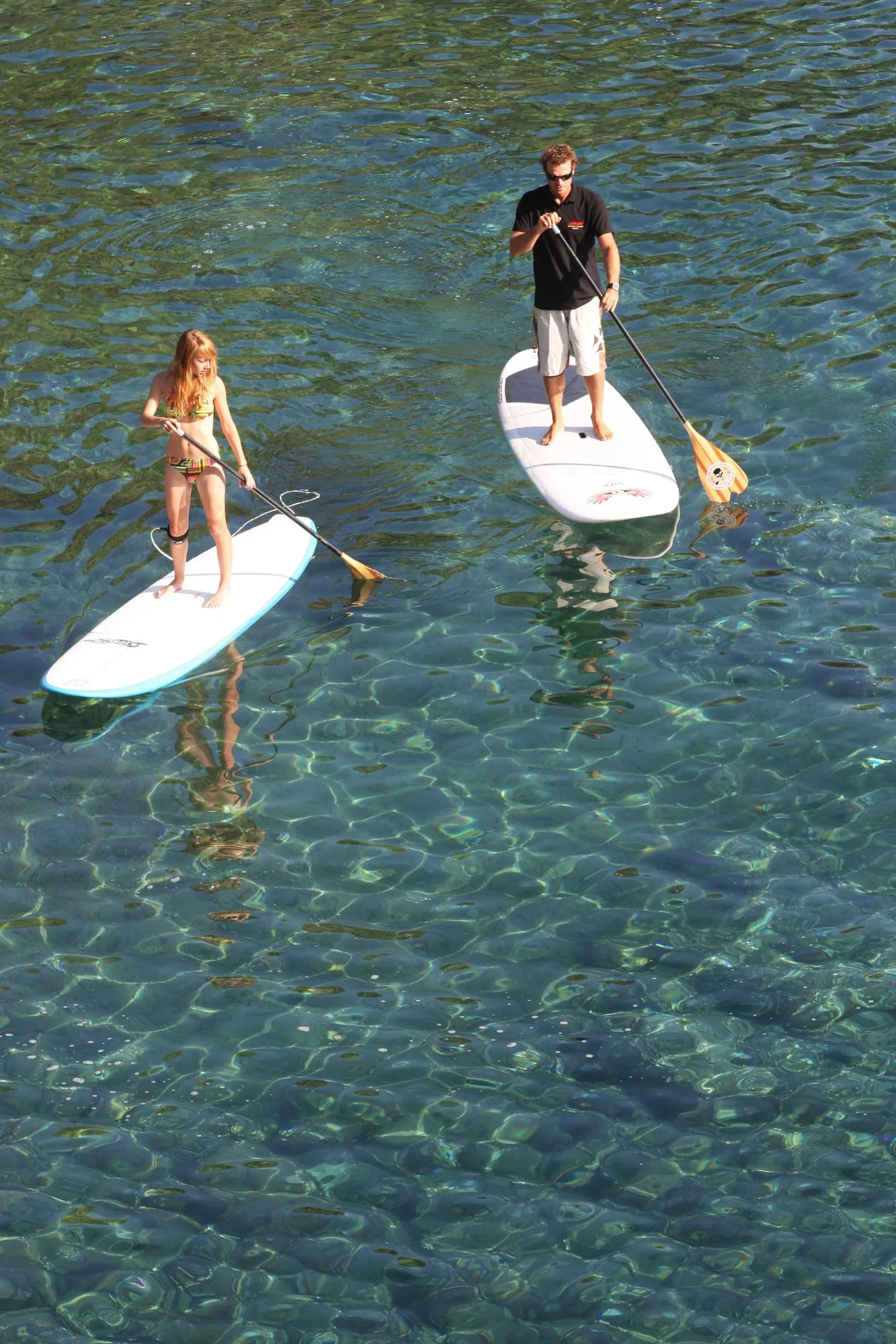 Dos personas remando de pie sobre tablas de paddle surf en agua transparente