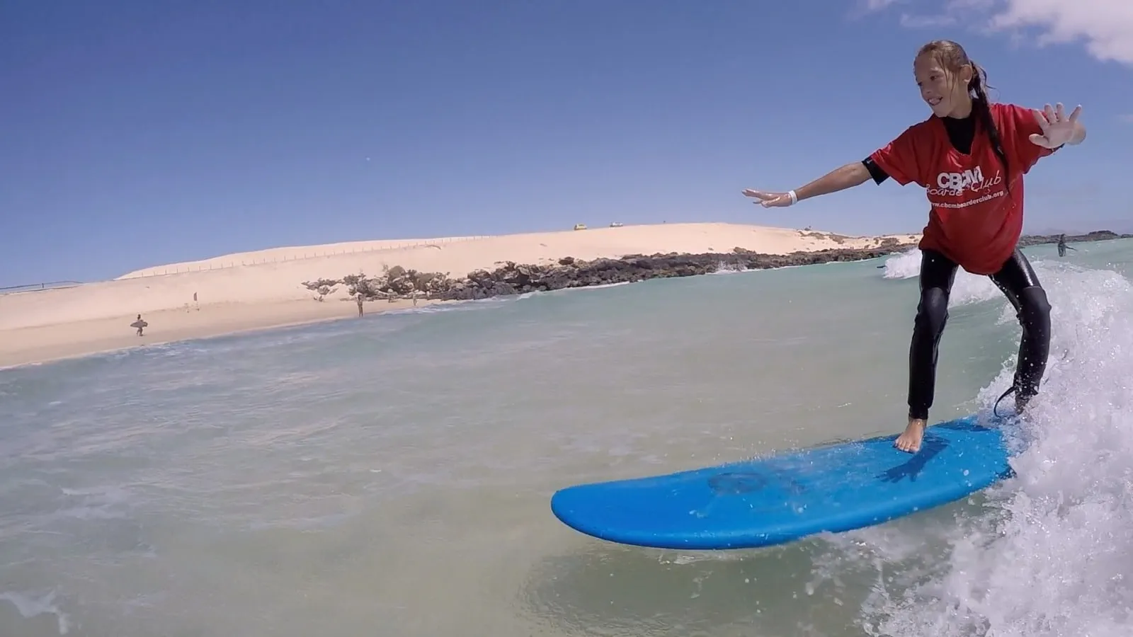 Alumna de CBCM de pie sobre una tabla azul, con las dunas de Corralejo al fondo