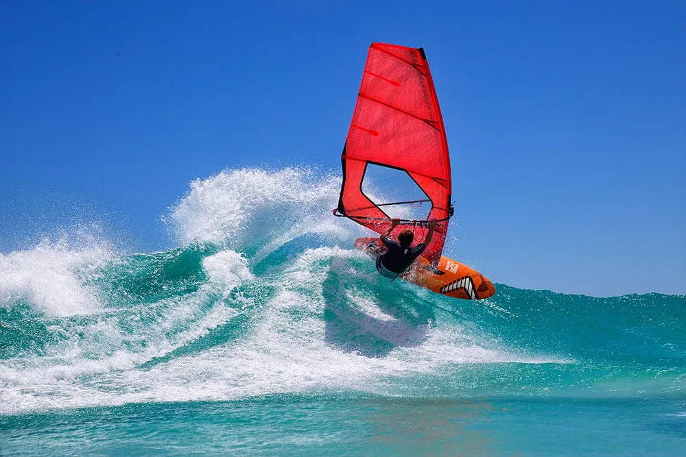 Windsurfista girando sobre la cresta de una ola con vela roja